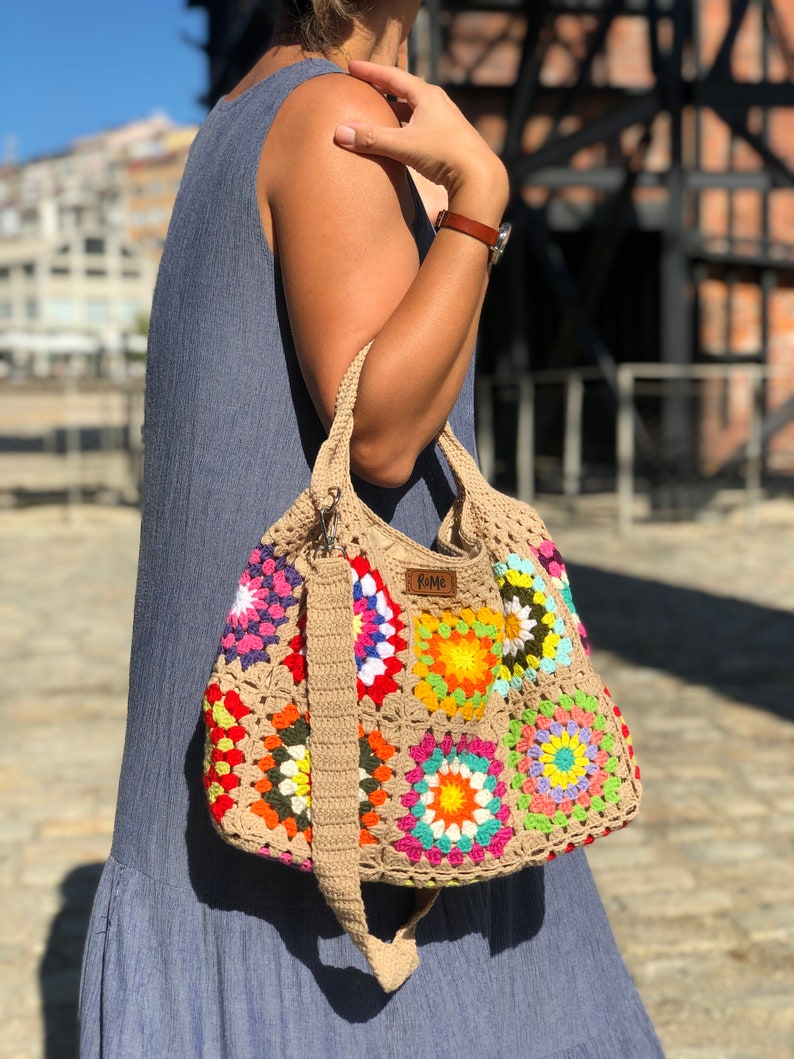 May include: A colorful, crocheted tote bag with a patchwork design. The bag features squares of various colors, including pink, yellow, orange, and blue, with a beige strap and handle. The bag has a label that reads "Rome".