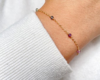 May include: Delicate gold chain bracelet adorned with small, colorful gemstones. The bracelet features a series of round, polished stones in shades of purple, pink, and green, spaced along the fine gold chain. The bracelet is shown on a wrist, against a white background.