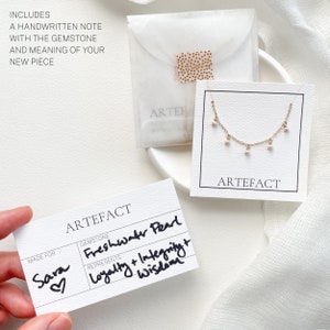 May include: A white card with the text "ARTEFACT" printed in black. The card has a section for "GEMSTONE" and "REPRESENTS". The gemstone is "Freshwater Pearl" and it represents "Loyalty + Integrity + Wisdom". The card also has a section for "MADE FOR" and the name "Sara" is written in cursive with a heart.