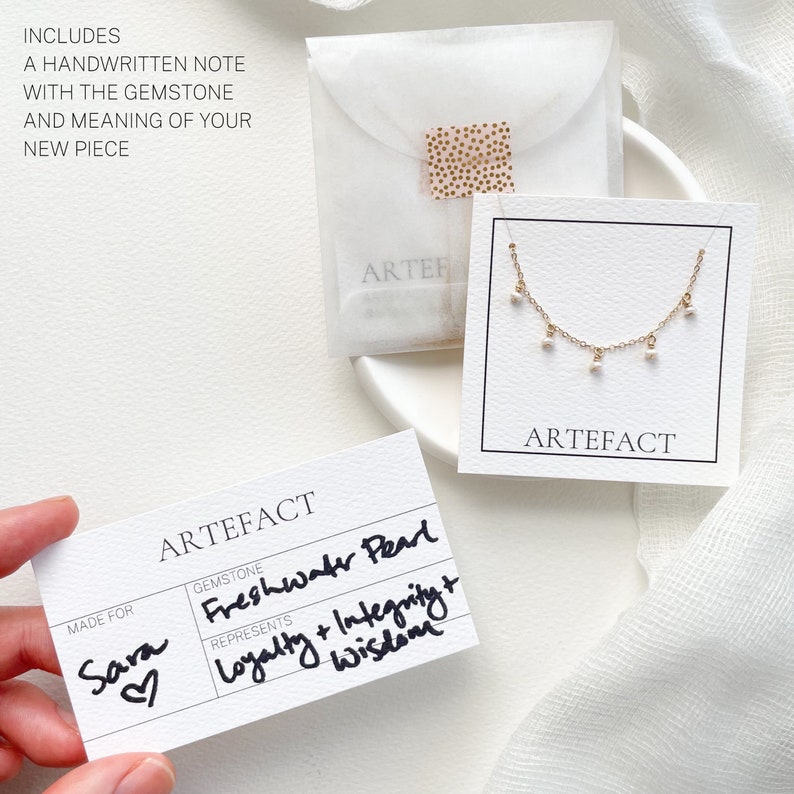 May include: A white card with the text "ARTEFACT" printed in black. The card has a section for "GEMSTONE" and "REPRESENTS". The gemstone is "Freshwater Pearl" and it represents "Loyalty + Integrity + Wisdom". The card also has a section for "MADE FOR" and the name "Sara" is written in cursive with a heart.