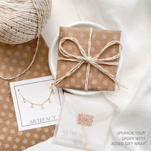 May include: A delicate gold necklace with small white pearls hanging from it. The necklace is displayed on a white card with the word "ARTEFACT" printed on it. The necklace is packaged in a brown paper gift bag with a white polka dot pattern and tied with a white twine bow. The text "UPGRADE YOUR ORDER WITH ADDED GIFT WRAP!" is printed on a white paper.