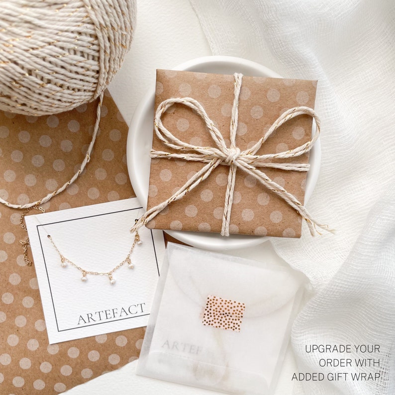 May include: A delicate gold necklace with small white pearls hanging from it. The necklace is displayed on a white card with the word "ARTEFACT" printed on it. The necklace is packaged in a brown paper gift bag with a white polka dot pattern and tied with a white twine bow. The text "UPGRADE YOUR ORDER WITH ADDED GIFT WRAP!" is printed on a white paper.