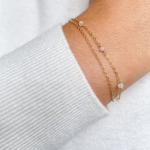 May include: A gold chain bracelet with small pink beads. The bracelet is layered on top of another gold chain bracelet.