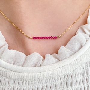 May include: A gold necklace with a bar pendant of small, faceted, ruby-coloured beads. The beads are strung horizontally, creating a minimalist design. The necklace is shown against a white background.
