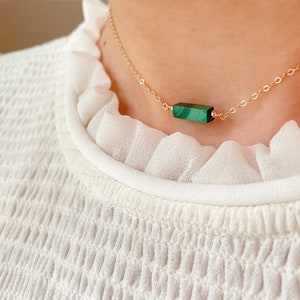 May include: A gold-colored chain necklace featuring a rectangular, green malachite stone pendant. The necklace is delicate and appears to be worn around the neck. The background is a white, textured fabric.