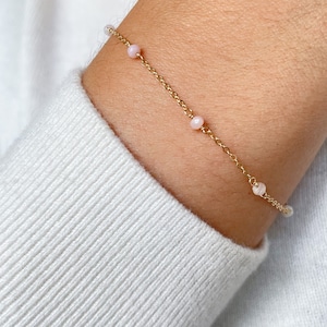 Peut inclure: Un bracelet délicat en chaîne dorée avec de petites perles roses. Le bracelet est porté au poignet.