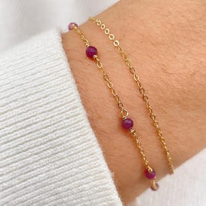 May include: Two gold chain bracelets with small, dark red gemstone beads. The bracelets are layered on a person's wrist.