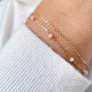 May include: A delicate gold chain bracelet with small pink opal beads. The bracelet is layered with a second, thinner chain.