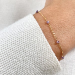 Peut inclure: Un bracelet en chaîne dorée avec de petites perles violettes. Le bracelet est porté au poignet.