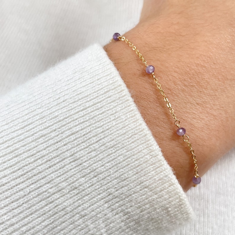 May include: A gold chain bracelet with small purple beads. The bracelet is on a person's wrist.
