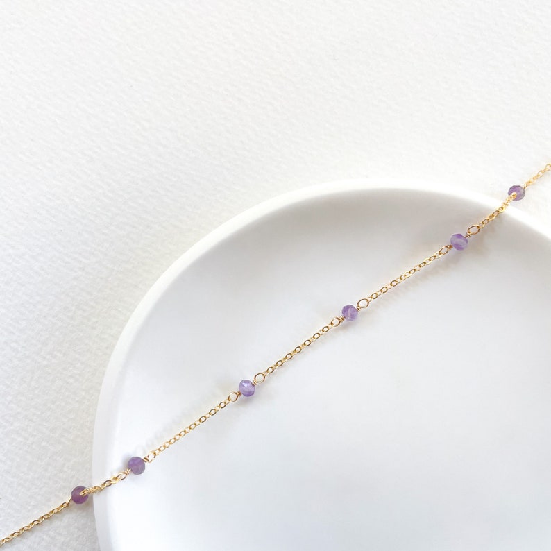 May include: A delicate gold chain necklace with small purple gemstone beads. The necklace is displayed on a white surface.