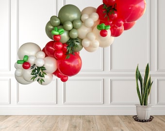 May include: A festive balloon garland in red, white, and green, with accents of faux berries and greenery. The balloons are arranged in a cascading design, perfect for a party or celebration. The garland is displayed against a white wall and wooden floor.