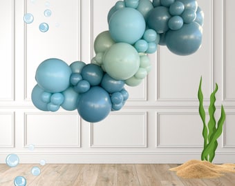 May include: A cluster of blue and green balloons, arranged in an arch, floats against a white wall. Bubbles and seaweed are also present, creating an underwater theme. The balloons vary in shades of blue and green.