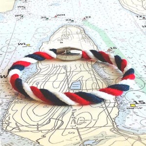 May include: A nautical-themed bracelet with a red, white, and blue twisted rope design. The bracelet features a silver-colored magnetic clasp. The background is a nautical chart.