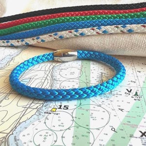 May include: A bright blue braided rope bracelet featuring a polished silver-tone metal magnetic clasp. The bracelet is circular in shape and constructed from a thick, woven cord material. Additional braided rope cords in black, red, green, and white are visible in the background.