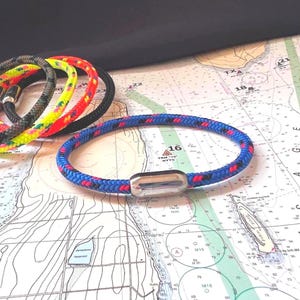 May include: A collection of nautical-themed bracelets in various colors, including blue, red, yellow, and black. Each bracelet features a braided rope design and a silver-tone magnetic clasp. The bracelets are displayed on a nautical chart.