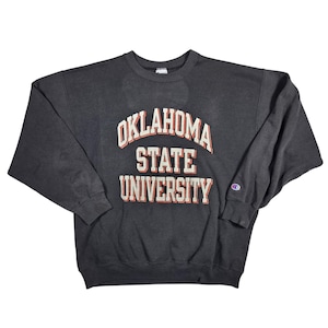 May include: Dark gray Oklahoma State University sweatshirt. The text "OKLAHOMA STATE UNIVERSITY" is printed in white with red accents. The Champion logo is visible on the left sleeve. The sweatshirt is a crew neck style.