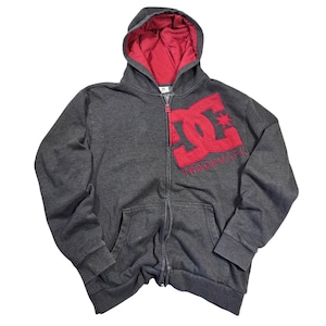 May include: A dark charcoal gray zip-up hoodie featuring a large red DC logo and the word TRADEMARK embroidered on the left chest. The garment includes a hood with a red interior lining, long sleeves with ribbed cuffs, and two front pockets.
