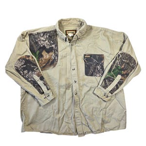 May include: A long-sleeve button-down shirt in tan with camouflage patterned panels on the shoulders and forearms. The shirt features a single chest pocket with a camouflage print and a Remington brand label on the collar and pocket.
