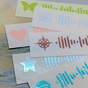 May include: Five different stickers with various designs and holographic finishes. The stickers feature a butterfly, a compass, a star, a steering wheel, and a Spotify logo. Each sticker has a corresponding Spotify barcode.