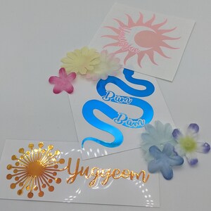 May include: Three vinyl stickers with different designs. The first sticker is a pink sun with a crescent moon and the text "Youngjae". The second sticker is a blue snake with the text "Pam Pam". The third sticker is a gold dandelion with the text "Yuggeom".
