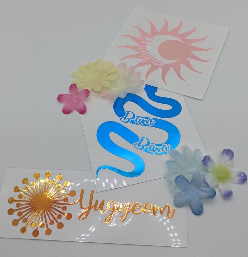 May include: Three vinyl stickers with different designs. The first sticker is a pink sun with a crescent moon and the text "Youngjae". The second sticker is a blue snake with the text "Pam Pam". The third sticker is a gold dandelion with the text "Yuggeom".