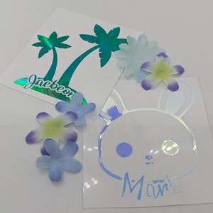 May include: Two iridescent vinyl stickers. One features a pair of palm trees with the text "Jaebeon" below. The other features a cartoon bunny with the text "Mavi" below.