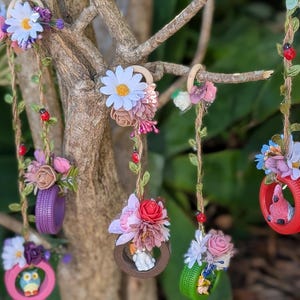 May include: A selection of miniature, brightly coloured tyre swings decorated with artificial flowers and ladybug details, suspended from a tree branch. The swings are painted in various colours, including red, purple, and green, and are decorated with a variety of flowers.