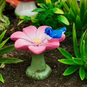 Puede incluir: Un bebedero de pájaros en forma de flor rosa con un pájaro azul posado en el borde. El bebedero está sentado en un lecho de hierba verde.