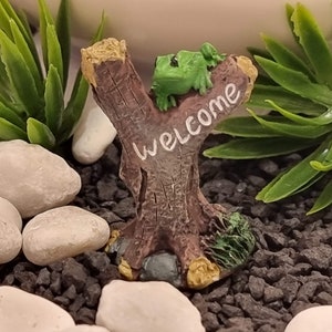 Puede incluir: Adorno de jardín en miniatura con una rana verde posada en un tronco de árbol marrón con la palabra "WELCOME" en blanco. La escena incluye piedras blancas, grava negra y plantas artificiales verdes.