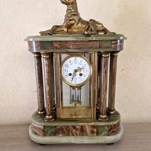 May include: A mantle clock featuring a bronze-toned dog figurine on top, supported by four green and brown marble-patterned columns. The clock face is white with black numerals and gold hands, housed in a glass case with a pendulum and a metal key.