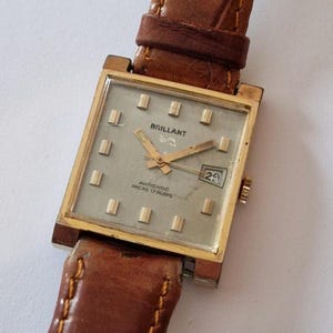 Może przedstawiać: Zegarek na rękę w stylu vintage w kolorze złotym z kwadratową tarczą i brązowym skórzanym paskiem. Tarcza zegarka jest jasnoszara ze złotymi znacznikami i wskazówkami. Zegarek wskazuje "BRILLANT" i "ANTICHOC ANCRE 17 RUBIS". Data ustawiona na 29.