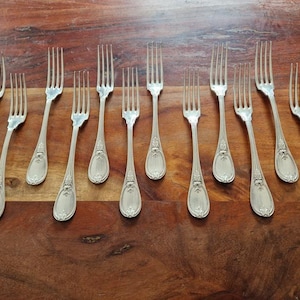 May include: A collection of silver forks with ornate detailing, arranged on a dark wooden surface. The forks have a classic design with a decorative pattern on the handles and tines. The arrangement suggests a formal dining setting.