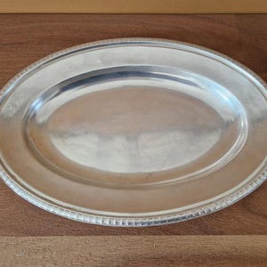May include: Oval silver-colored serving platter with a beaded edge. The platter is made of metal and has a smooth, reflective surface. The serving dish is suitable for serving food at a dinner party or special event.