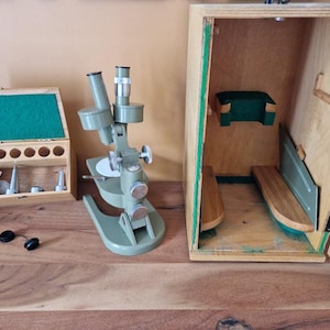 Op de afbeelding: Een vintage olijfgroene microscoop met een houten opbergdoos. De doos is open en onthult compartimenten en accessoires. Een aparte houten koffer met een groen viltinterieur bevat extra componenten. Zwarte oculairs zijn ook zichtbaar.
