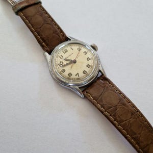 Puede incluir: Reloj de pulsera plateado vintage con esfera crema y números negros. El reloj, con la inscripción "ELVETIA", tiene una correa de cuero marrón con un patrón texturizado. Un reloj clásico.