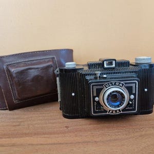 May include: A black and silver vintage camera with a leather case. The camera has a lens with the text "Ultra Fex" and "Automatic Speed" printed on it.