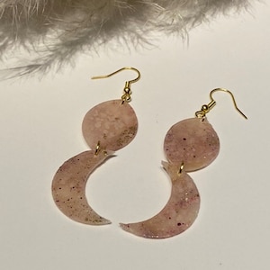 Könnte beinhalten: Ohrringe mit Mondphasen-Design. Jeder Ohrring hat einen goldfarbenen Haken, einen kreisförmigen oberen Teil und eine Halbmondform. Die Ohrringe sind hellrosa mit goldenen und violetten Sprenkeln. Das Thema ist himmlisch.