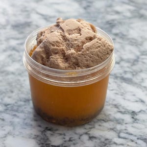 Könnte beinhalten: Ein durchsichtiger Plastikbehälter gefüllt mit einer braunen, geschlagenen Creme und einer Schicht orangefarbener Flüssigkeit.