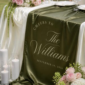 May include: A dark olive green satin table runner draped over a white tablecloth, featuring the white printed text "CHEERS TO The Williams SEPTEMBER 19, 2026" in a mix of serif and script fonts, creating a smooth, reflective surface texture.