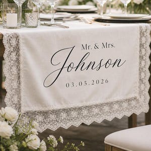 May include: A white fabric table runner featuring intricate lace trim along the edges. The center is printed with the black text Mr. & Mrs. Johnson and the date 03.05.2026 in a combination of serif and elegant script fonts on the smooth surface.