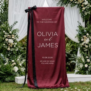 May include: A tall, rectangular, deep burgundy fabric wedding welcome sign featuring white text that reads: WELCOME TO THE WEDDING OF OLIVIA and JAMES 10.08.2026 WE ARE SO GLAD YOU ARE HERE! A long, black satin ribbon is tied in a bow at the top left corner.