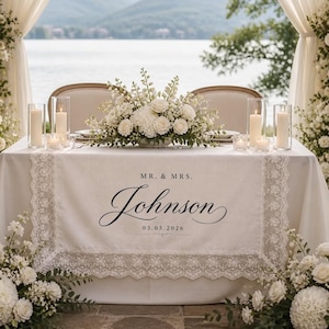 May include: A white wedding table runner featuring lace trim and the printed text MR. & MRS. Johnson 03.05.2026. The runner is displayed on a table with a floral centerpiece, candles, and white curtains, set against a backdrop of a lake and mountains.