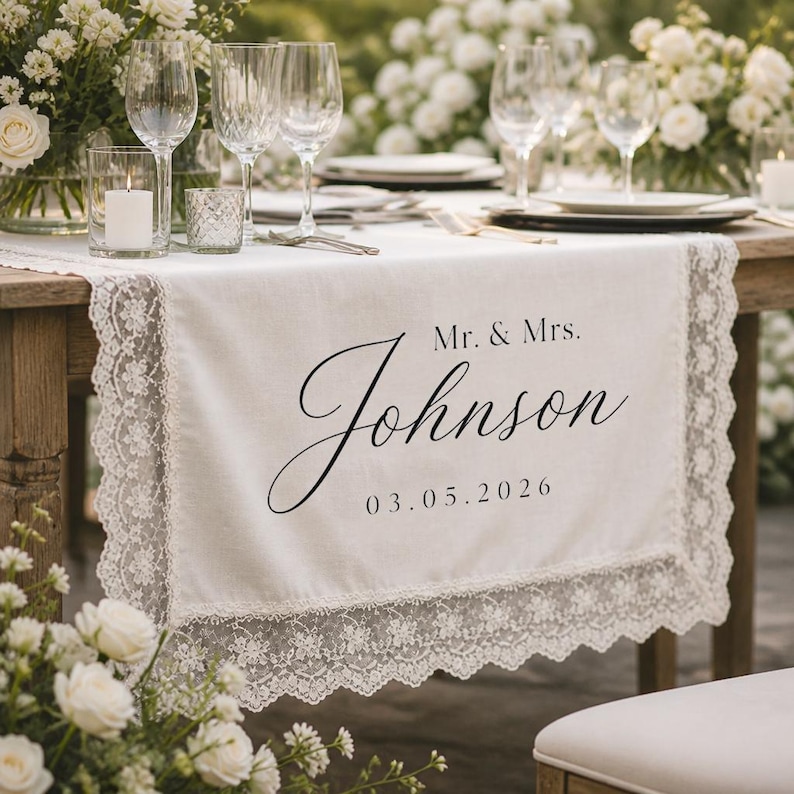 May include: A white fabric table runner featuring intricate lace trim along the edges. The center is printed with the black text Mr. & Mrs. Johnson and the date 03.05.2026 in a combination of serif and elegant script fonts on the smooth surface.