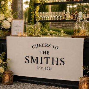 May include: A cream-colored fabric banner featuring the black printed text CHEERS TO THE SMITHS EST. 2026. The banner is displayed on the front of a bar setup with glass bottles, glassware, a framed menu, and lanterns placed on the counter surface.