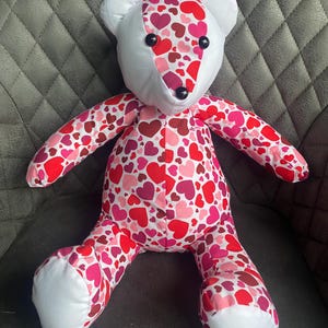 May include: A plush teddy bear with a white face and pink and red heart pattern on its body. The bear is sitting on a gray textured surface.