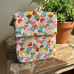 May include: A fabric tote bag with a flap closure, patterned with colourful cupcakes on a white background. The cupcakes feature various frosting and topping designs. The bag is on a distressed wooden surface.