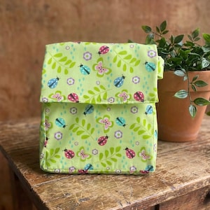 May include: A lime green fabric storage container with a flap, decorated with a pattern of ladybugs, butterflies, flowers, and leaves. The container is sitting on a wooden surface, with a potted plant in the background.