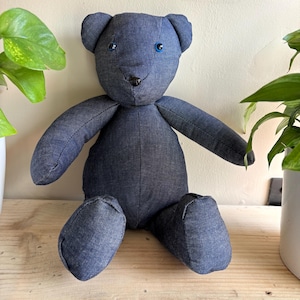 May include: A denim teddy bear with blue eyes and a black nose sits on a wooden surface. The bear's arms and legs are extended, and it has a classic teddy bear shape. The bear is surrounded by green plants.