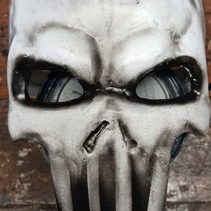 May include: A white skull mask with black stripes and eye holes. The mask is made of a smooth, hard material.